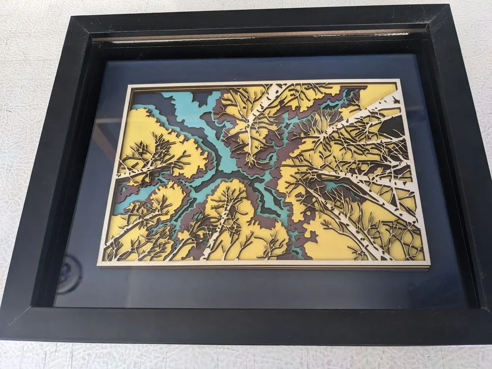 Layered laser-cut river and forest scene in yellow and teal, black frame