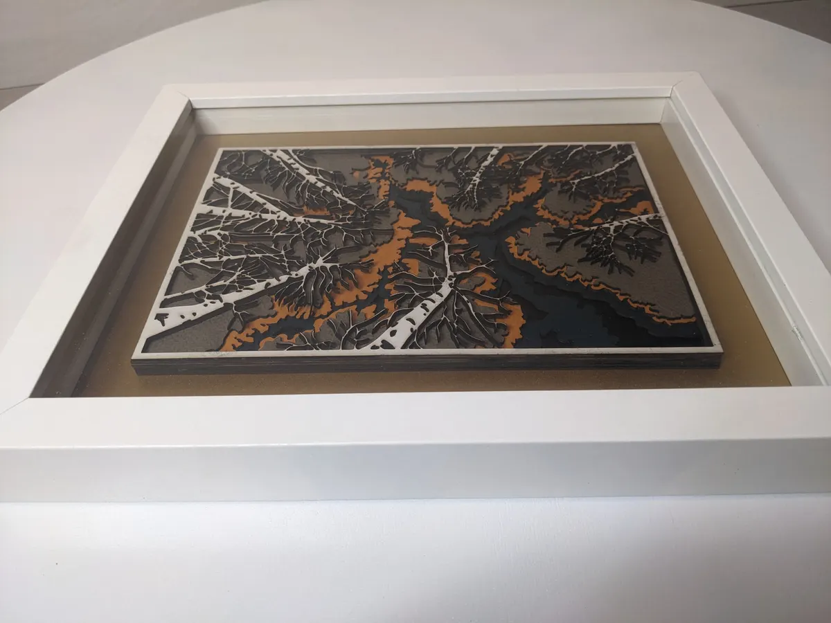 Layered laser-cut river scene in orange and grey in a white shadow box